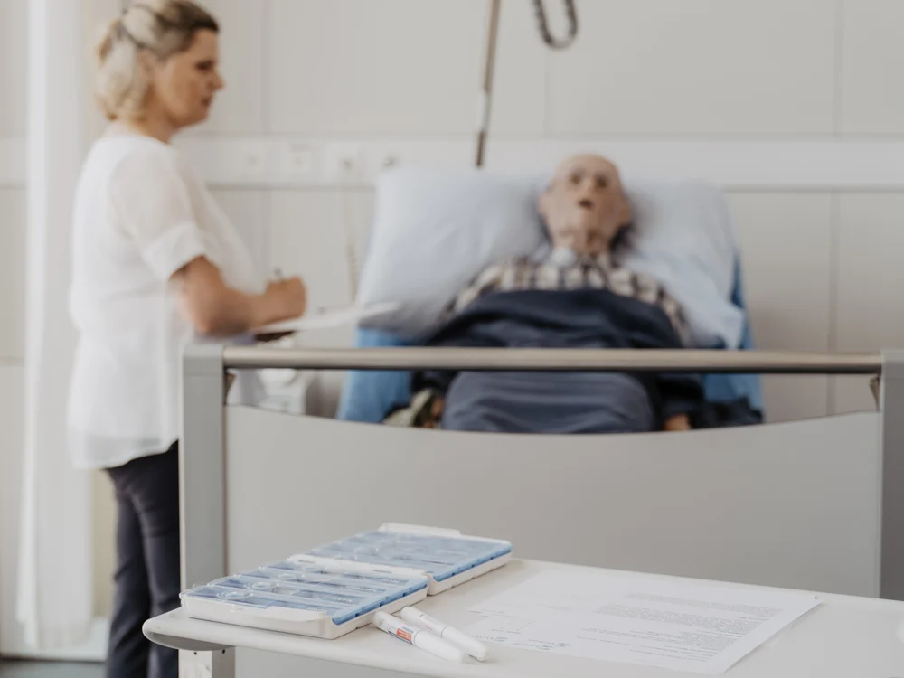 Material auf einem Tisch; dahinter eine Teilnehmerin und die Pflegepuppe im Bett.